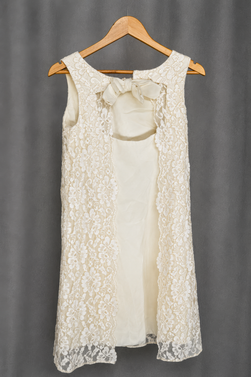Sleeveless ivory dress on a wooden hanger. Two vertical floral lace panels run down the front over a solid ivory center panel, with the center open between the lace panels. A fabric bow sits at the neckline above the opening.