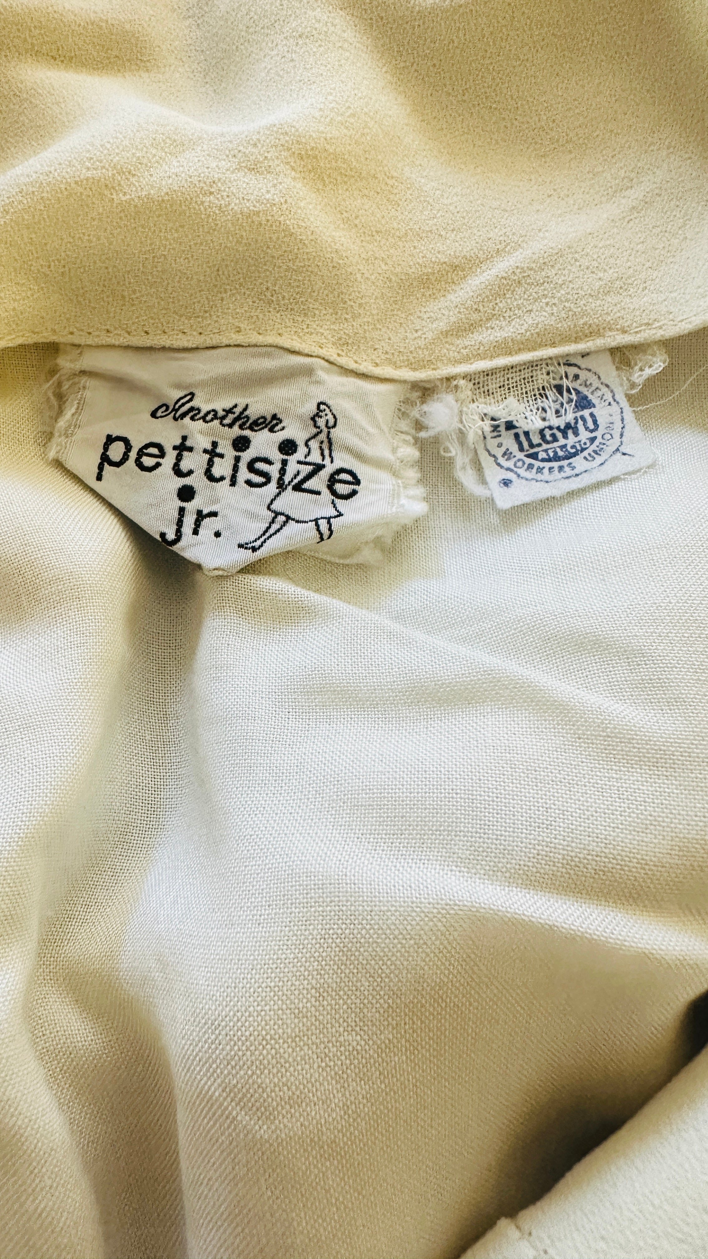 Interior fabric tag with cursive "Another" above block letters reading "pettisize jr.", printed with a small figure of a woman. Positioned at the garment's inner neckline.ILGWU / International Ladies' Garment Workers' Union".