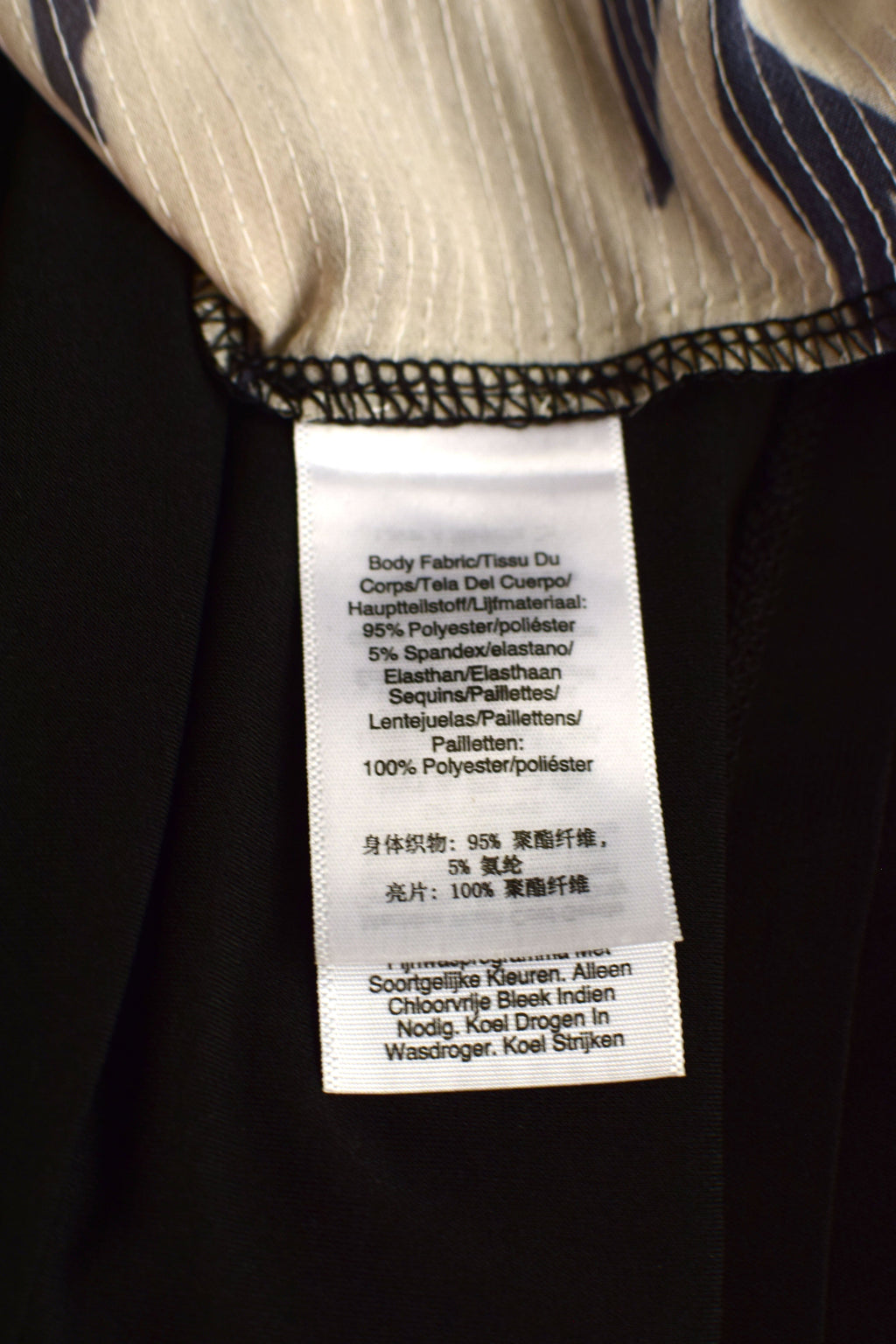 Close-up of a garment's interior label sewn onto a black fabric body, with a striped inner lining visible at the top. The white tag contains product details in Dutch and English.
