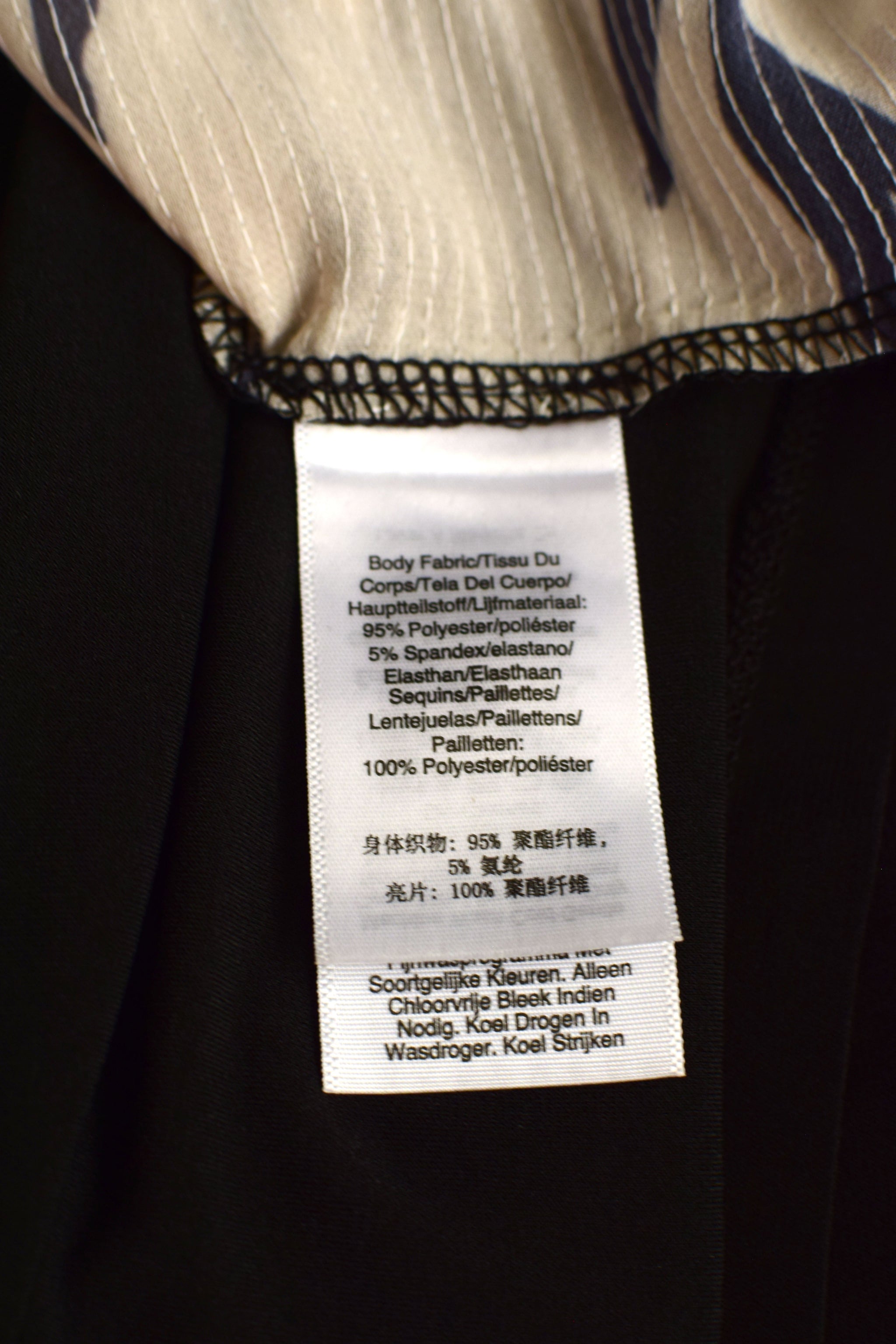 Close-up of a garment's interior label sewn onto a black fabric body, with a striped inner lining visible at the top. The white tag contains product details in Dutch and English.