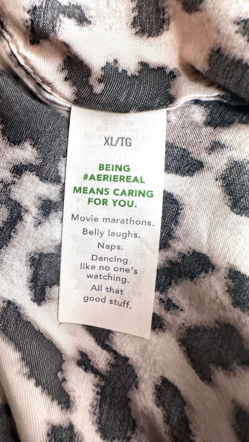 Close-up of a garment tag reading 'XL/TG' and 'BEING #AERIEREAL MEANS CARING FOR YOU.' with additional text, placed on a black-and-white patterned fabric background.
